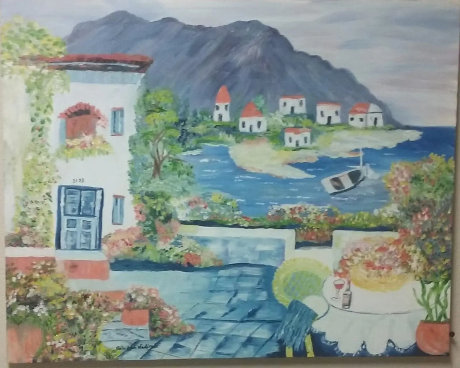 Painted coastal village with white buildings, a dark mountain, blue water, a small boat, flowers and a terrace table.